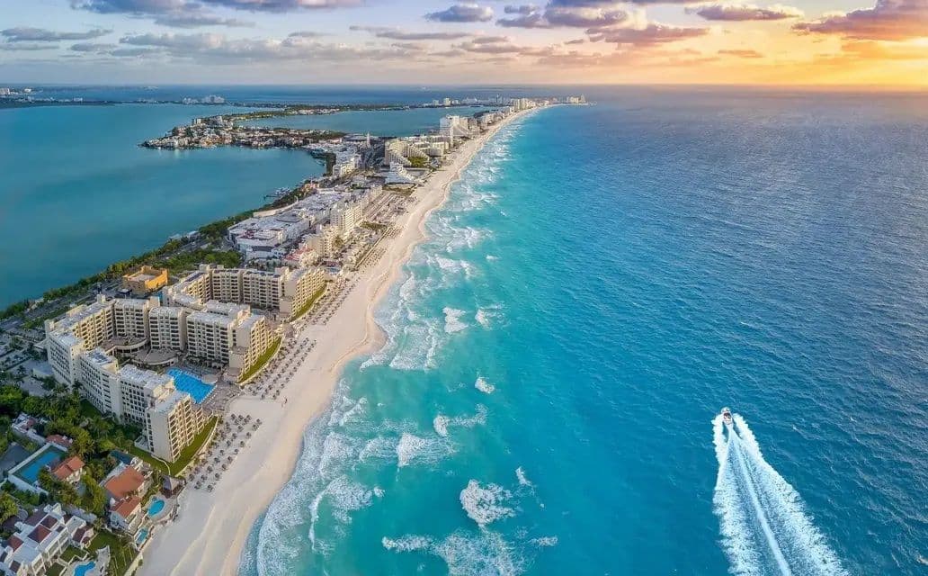 Vista aerea di una costa con hotel e resort su una spiaggia di sabbia bianca e acque turchesi, con un motoscafo che lascia una scia.