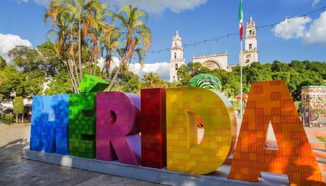 Grandi lettere colorate che compongono 'MERIDA' si ergono in una piazza con una chiesa, palme e una bandiera messicana sullo sfondo.