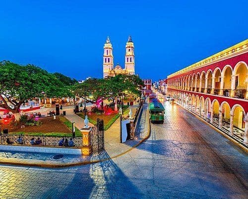 Una veduta dall'alto di una piazza cittadina al crepuscolo, che mostra una cattedrale illuminata, un parco e un tram verde su una strada acciottolata.