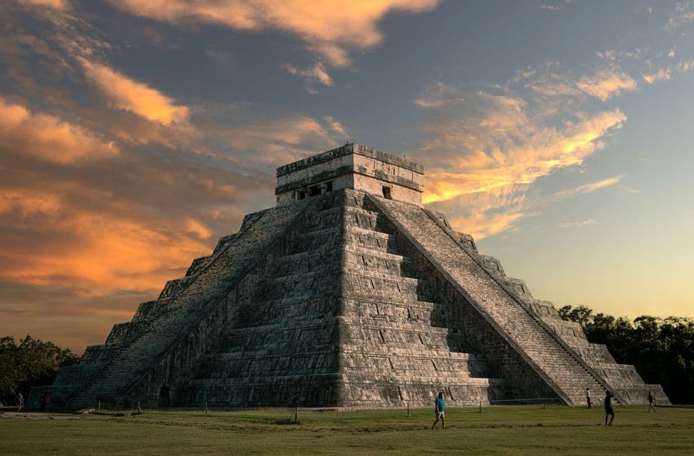 Una grande e antica piramide a gradoni in pietra, con una ripida scalinata, si erge contro un cielo pieno di nuvole arancioni al tramonto.