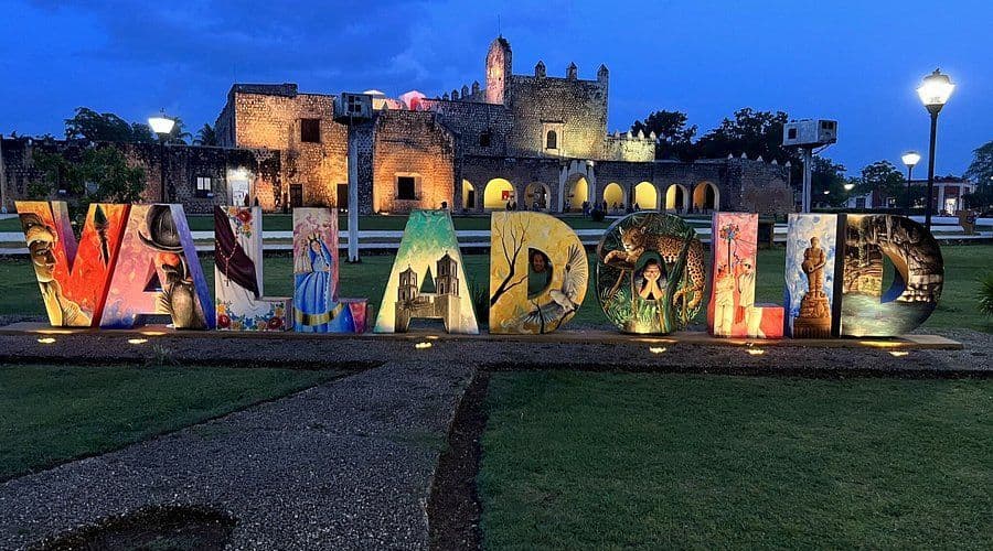 Grandi lettere colorate e illuminate che compongono 'Valladolid' su un prato al crepuscolo, con un vecchio edificio in pietra illuminato sullo sfondo.