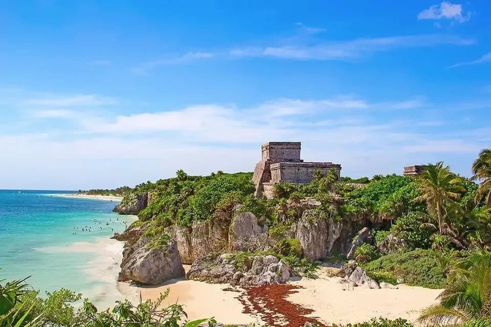 Una rovina di un tempio in pietra si erge su una scogliera verde e rocciosa, sopra una spiaggia sabbiosa e un oceano turchese.