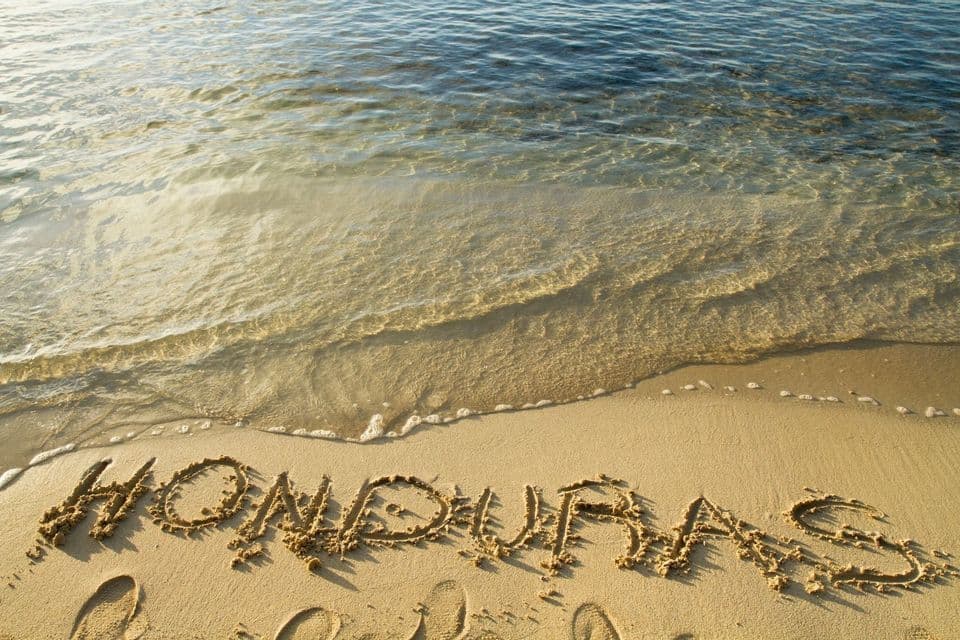 Das Wort Honduras im Sand an einem Strand geschrieben, wobei klares Meerwasser sanft an das Ufer spült.