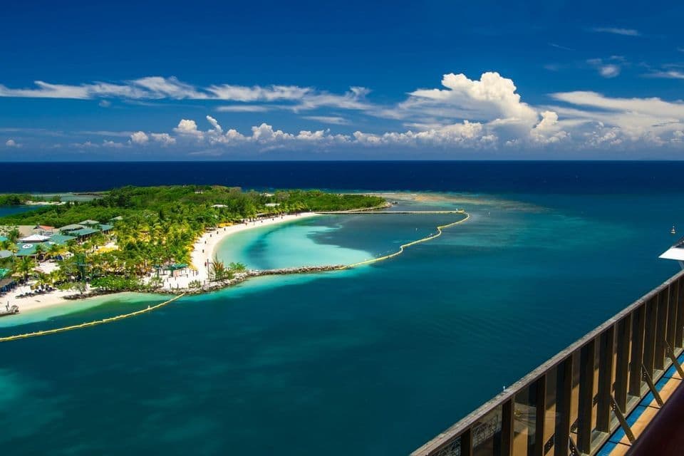 Luftaufnahme einer tropischen Insel mit einem weißen Sandstrand, der an eine ruhige, türkise Bucht grenzt, unter einem strahlend blauen, teilweise bewölkten Himmel.