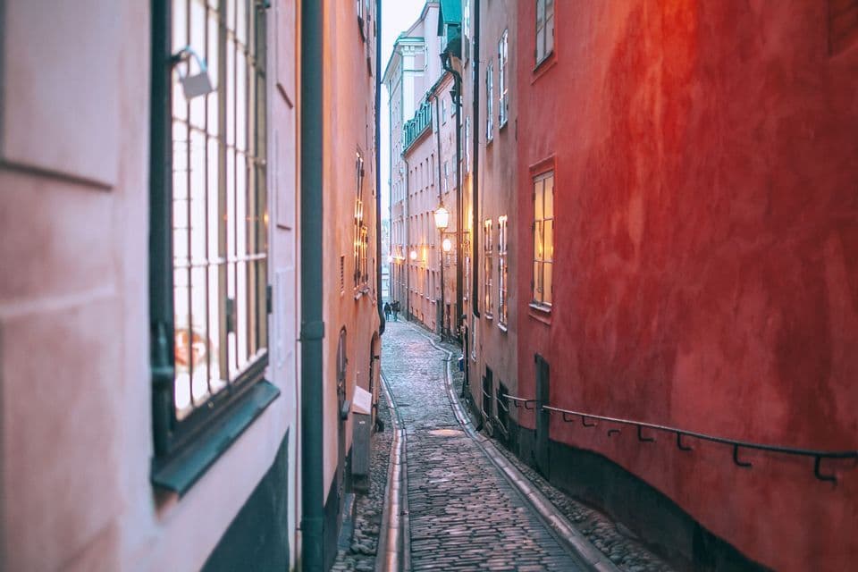 A narrow cobblestone alley winds between tall, colorful buildings, illuminated by street lamps at dusk.