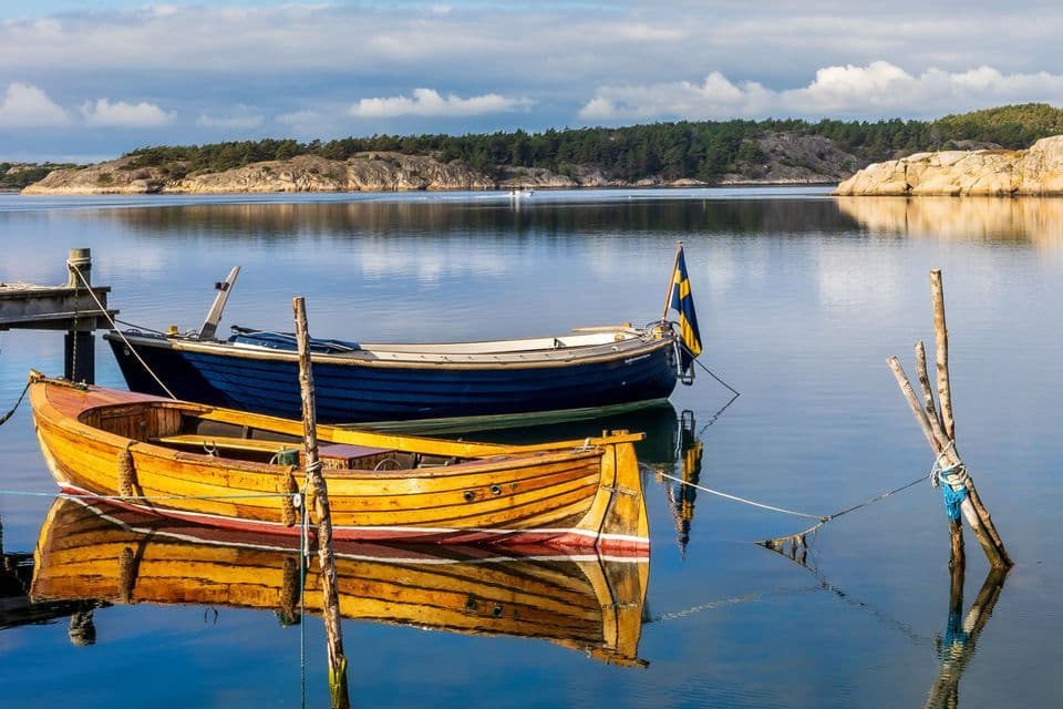 Two wooden boats, one yellow and one blue with a Swedish flag, are moored in calm water with their reflections on the surface.
