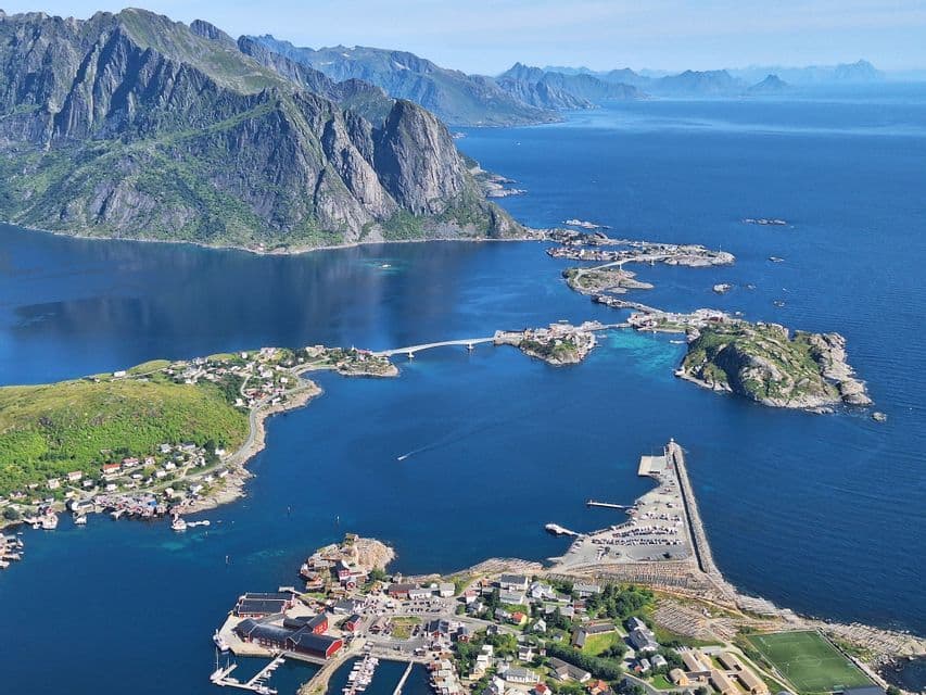 An aerial view of a coastal village on small islands connected by bridges, with a dramatic mountain range rising from the blue sea.