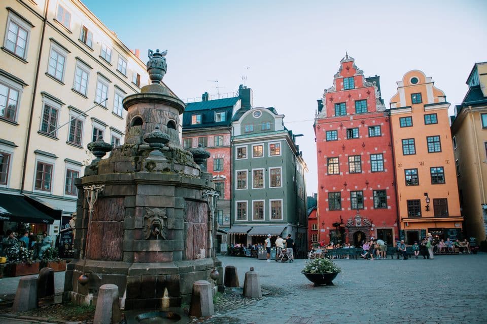 An ornate stone fountain sits in a cobblestone square, surrounded by people and colorful, traditional European buildings.