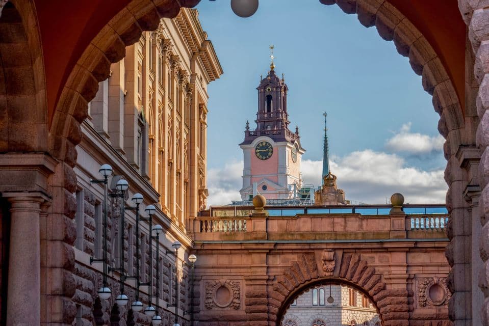 A pink clock tower and a classical building are viewed through a large, ornate stone archway under a partly cloudy sky.