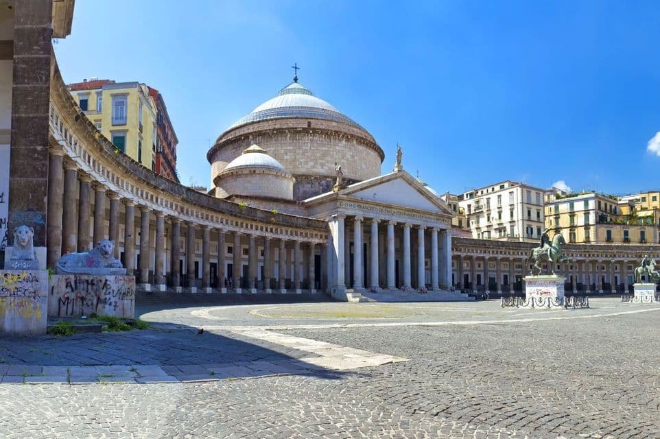 A domed church with a curved colonnade stands in a large, cobblestone piazza under a bright blue sky.