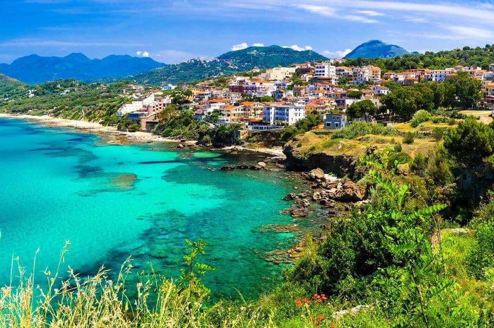 A colorful coastal village on a green hillside overlooks a clear turquoise sea with a rocky shore and distant mountains.