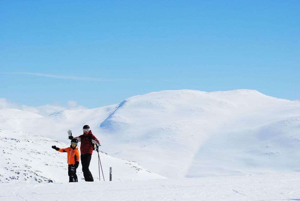 Due partecipanti a un viaggio di gruppo WeRoad, in tenuta da sci, in piedi su una montagna innevata, con uno che saluta, sotto un cielo azzurro e limpido.