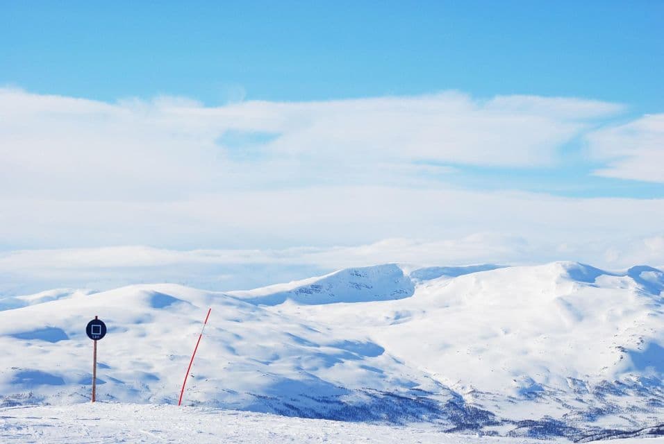 Un cartello blu di pista da sci su un pendio innevato che domina una vasta catena montuosa sotto un cielo blu.