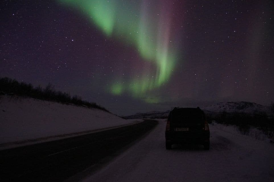 Un'auto scura è parcheggiata a lato di una strada innevata di notte, con l'Aurora Boreale verde e viola visibile nel cielo stellato.