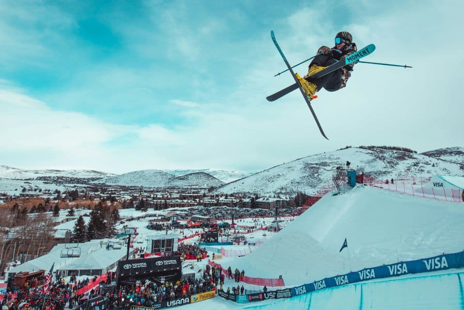 Un esquiador acrobatico realiza un truco aereo con los esquis cruzados sobre una rampa de nieve durante una competicion con una multitud abajo