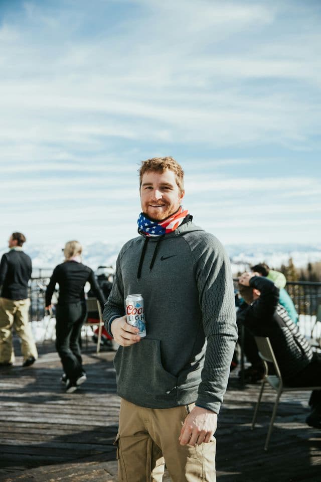 Un hombre en un viaje en grupo de WeRoad sonríe mientras sostiene una lata de cerveza en una terraza de madera en una zona montañosa nevada.