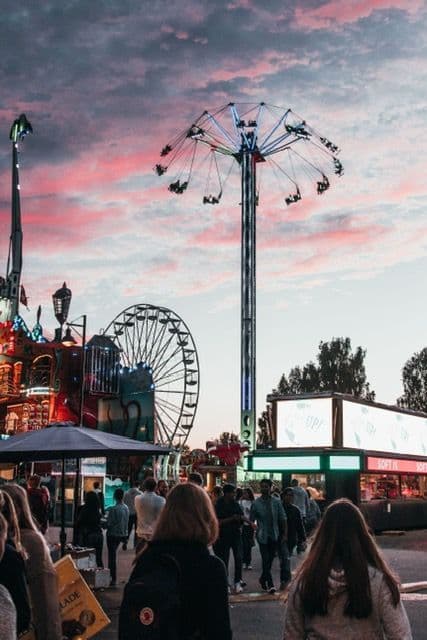 A crowd of people walks through an amusement park with a swing ride and a Ferris wheel operating against a pink sunset sky.