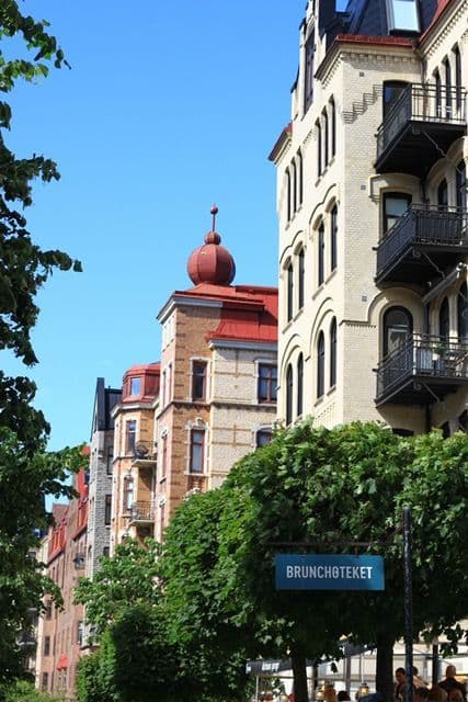 A street-level view of classic European apartment buildings with a red turret and green trees under a clear blue sky.