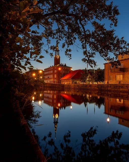 A brick building with a clock tower stands by a river at dusk, its reflection visible on the water's surface from under a tree.