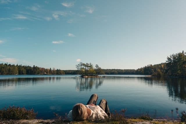 A person lies on their back on a rocky shore, looking out at a calm lake with a small, tree-covered island.