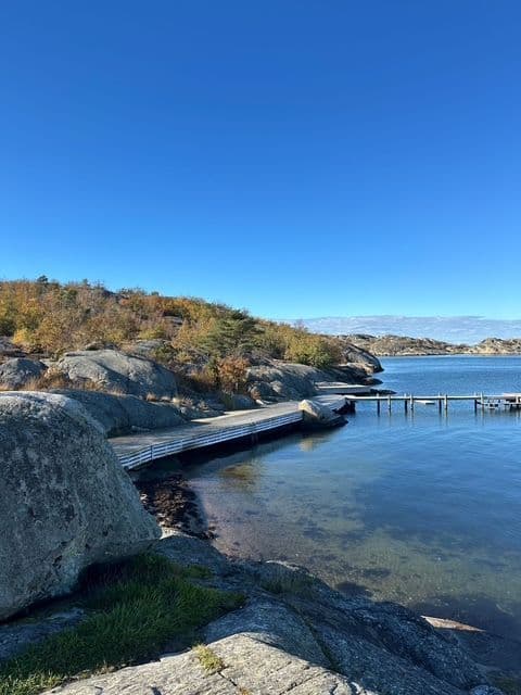 A wooden boardwalk curves along a rocky shoreline next to calm, clear water under a bright blue sky.