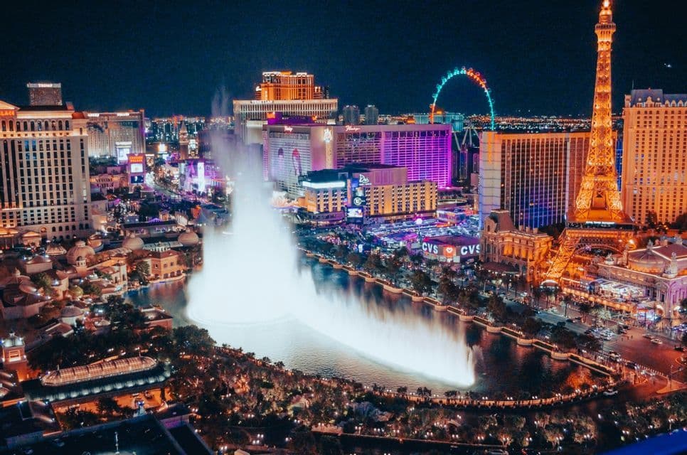 Vista aerea del vibrante horizonte de una ciudad por la noche, con grandes fuentes en erupción en un lago y una réplica iluminada de la Torre Eiffel.