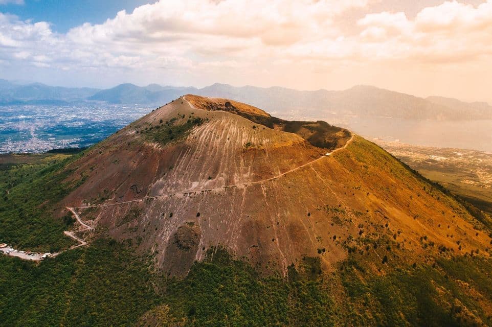 Una vista aérea de un gran cráter volcánico con un sendero a lo largo de su borde, con vistas a una ciudad costera bajo un cielo parcialmente nublado.