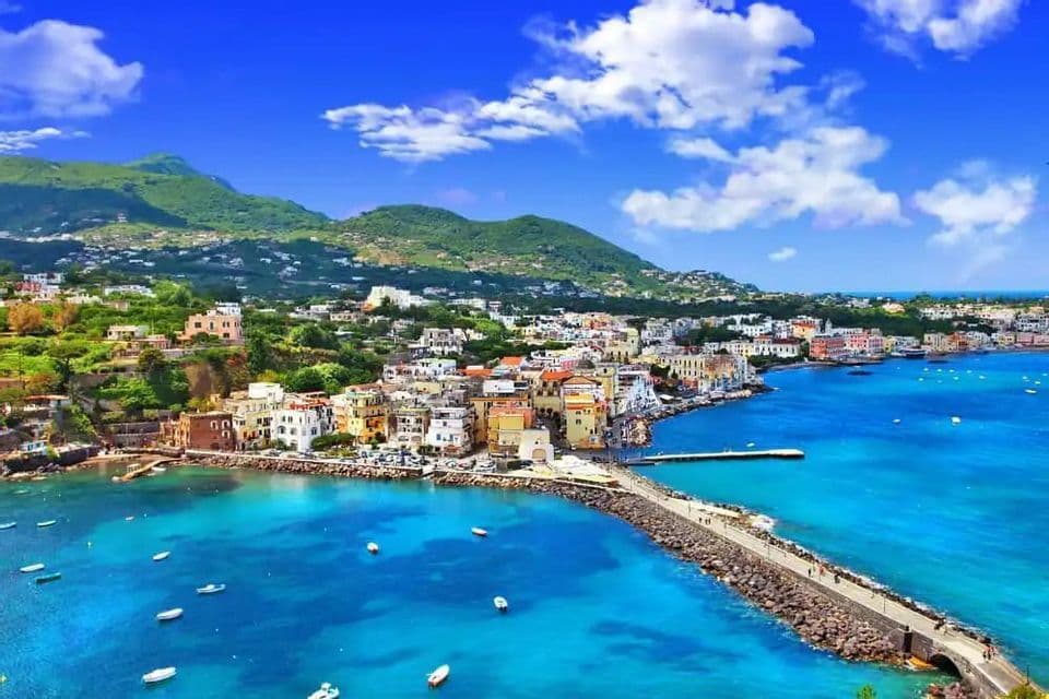 A colorful coastal town meets a turquoise sea with a stone pier, under green hills and a bright blue sky.