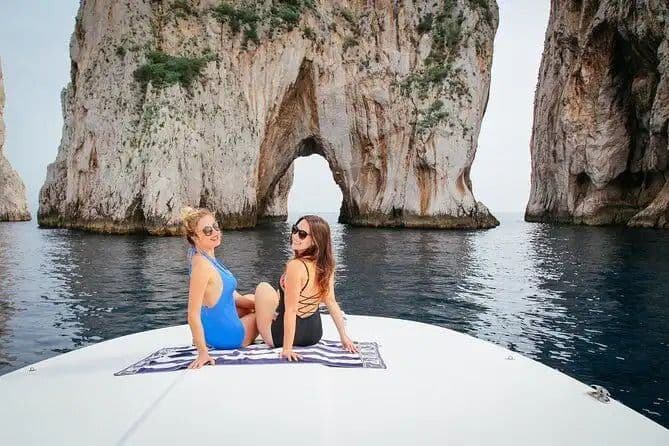 Two women in swimsuits sit on the bow of a boat, smiling in front of large sea stacks with a natural arch.