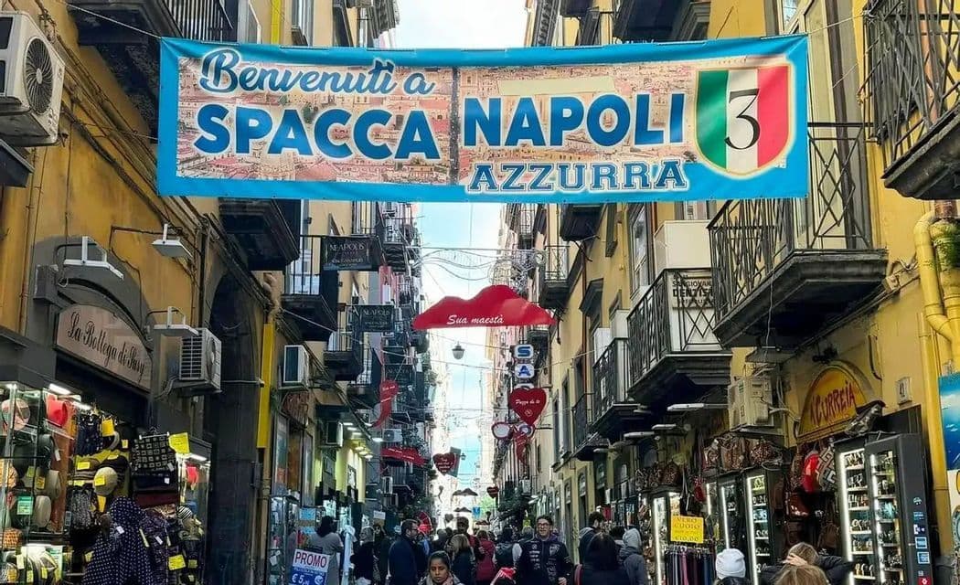 A large banner celebrating Napoli hangs over the crowded Spaccanapoli street, lined with shops and people.
