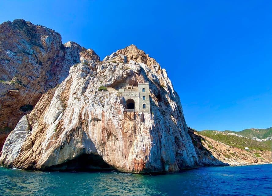 A stone building with an arched tunnel entrance built into a steep sea cliff above the blue ocean.