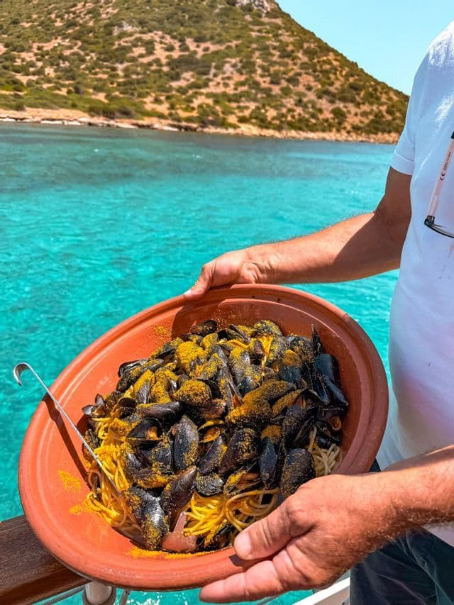 A person holds a large terracotta bowl of spaghetti with mussels, seasoned with yellow spices, over turquoise water.