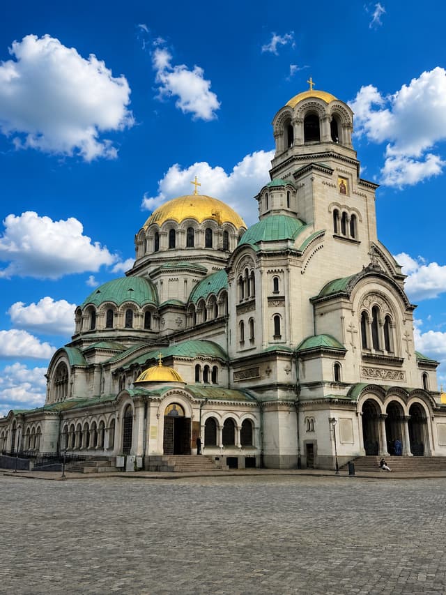 Une cathédrale de pierre ornée avec des dômes dorés et verts se dresse sur une place pavée sous un ciel bleu éclatant.