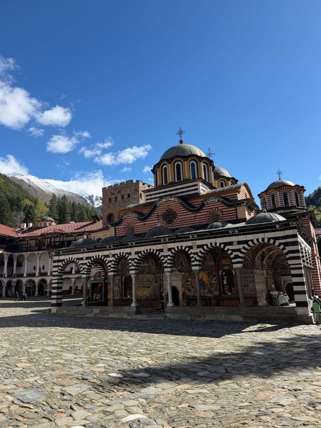 Un monastère orné d'arches rayées noires et blanches se dresse dans une cour pavée, sur fond de montagnes enneigées.