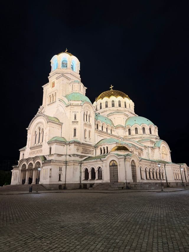 Une grande cathédrale en pierre blanche avec des dômes dorés et verts, illuminée la nuit sur un ciel sombre au-dessus d'une place pavée.