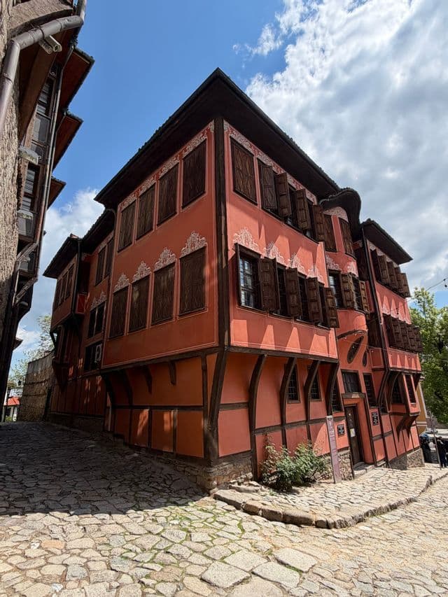 Vue en contre-plongée d'une maison traditionnelle couleur ocre avec des colombages et des volets marron foncé à l'angle d'une rue pavée.