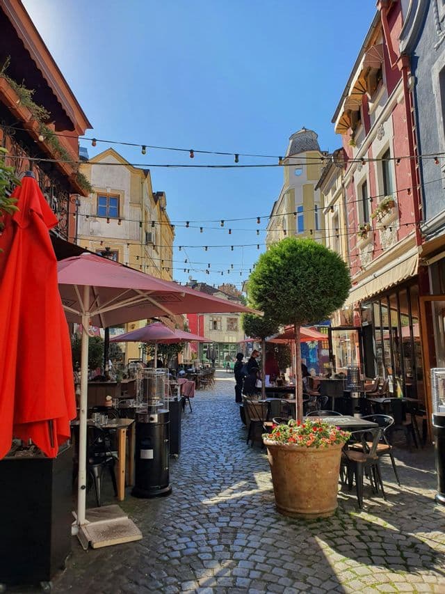 Une ruelle pavée ensoleillée et étroite, bordée de cafés en plein air, avec des tables, des parasols et des guirlandes lumineuses suspendues.