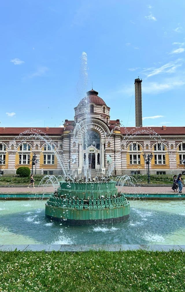 Une fontaine à étages verte projette de l'eau sur une place devant un bâtiment orné et rayé avec un dôme par une journée ensoleillée.