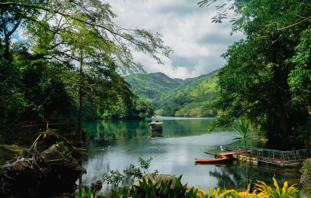 Un lac tranquille entouré d'arbres et de collines verdoyantes, avec une cabane flottante sur l'eau et un bateau orange près d'un quai.