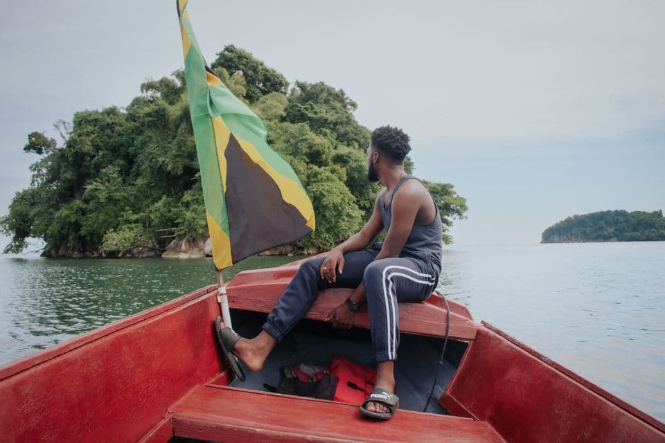 Un homme est assis sur le flanc d'un bateau rouge arborant un drapeau jamaïcain, contemplant une île verdoyante et luxuriante sur l'eau.