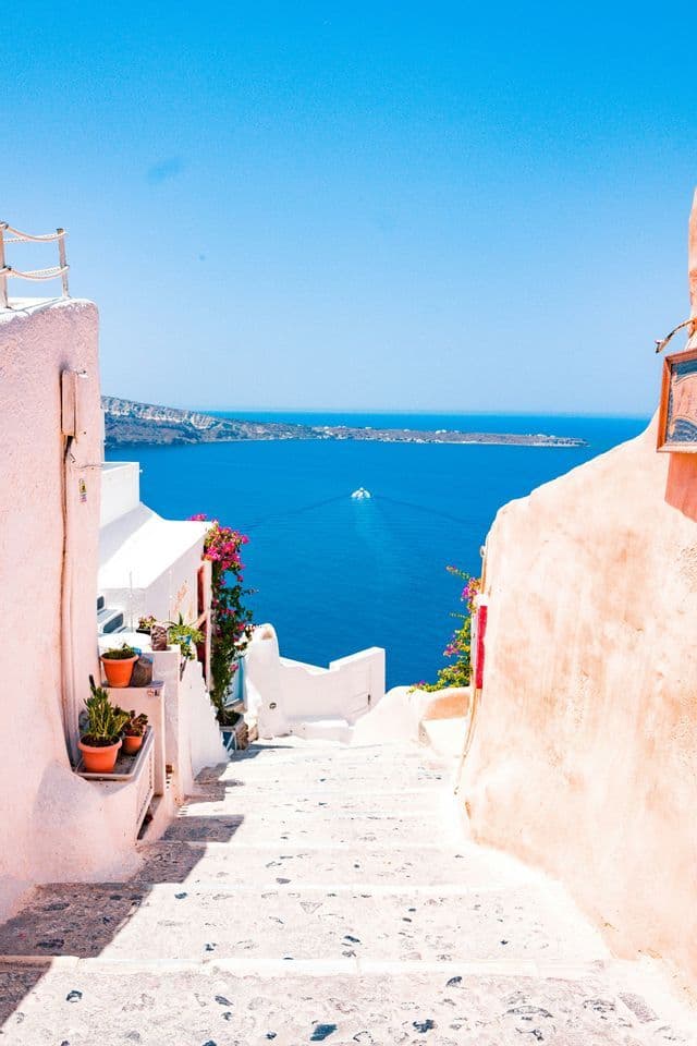 Eine schmale Steintreppe führt zwischen weiß getünchten Gebäuden hinunter und bietet Aussicht auf das blaue Meer und eine ferne Insel.