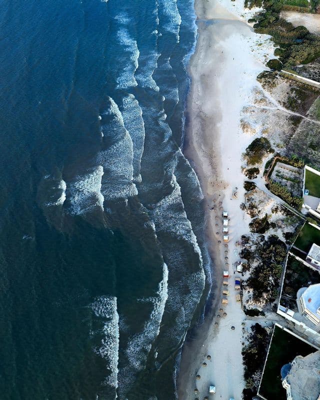 Eine Luftaufnahme von tiefblauen Meereswellen mit weißem Schaum, die an einen Sandstrand neben Küstengebäuden rollen.