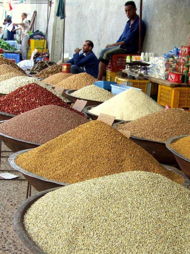 Auf einem Marktstand im Freien werden Stapel verschiedener Getreidesorten und Hülsenfrüchte in großen Schalen präsentiert, dahinter sitzen zwei Verkäufer.