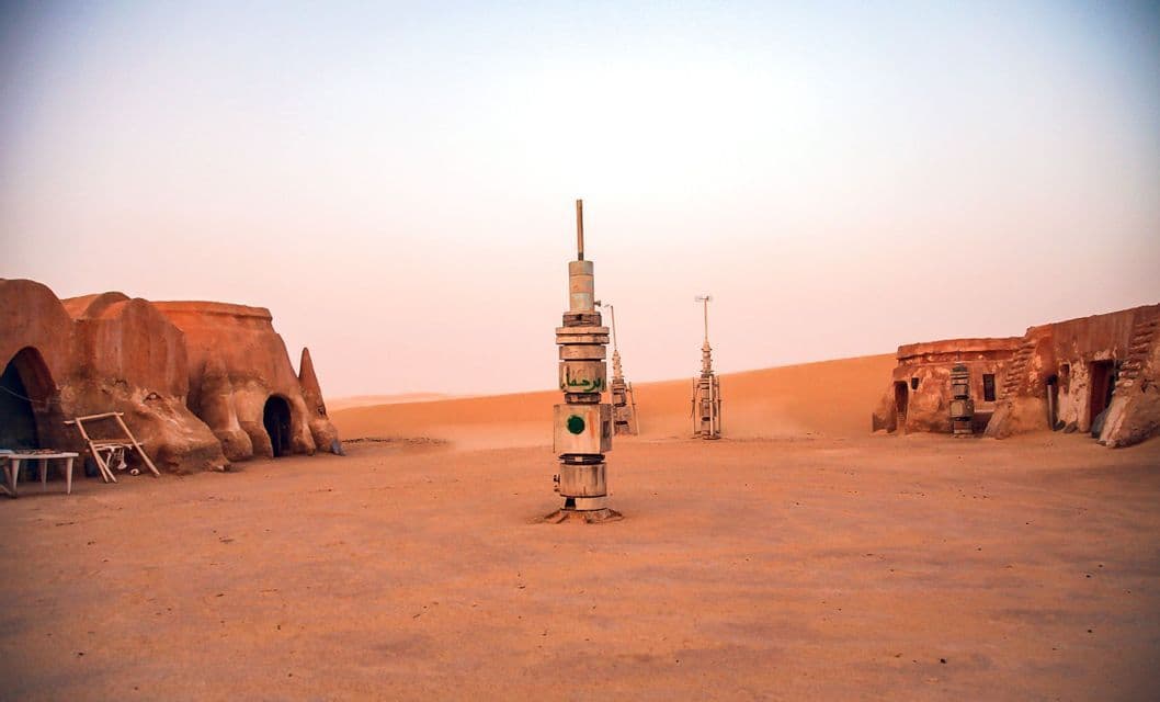 Futuristische Türme und Lehmziegelbauten stehen in einer Wüstenlandschaft unter einem fahlen Himmel in der Dämmerung.