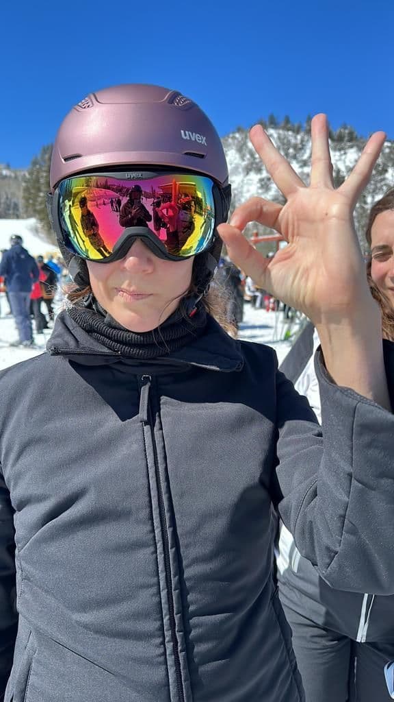 Una mujer en un viaje en grupo de WeRoad hace un gesto de aprobación con la mano, vistiendo un casco de esquí y gafas reflectantes en una pista de montaña soleada y nevada.