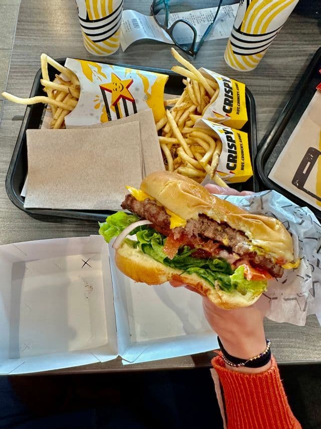 La mano de una persona sostiene una hamburguesa parcialmente comida sobre una mesa con una bandeja de papas fritas y bebidas.