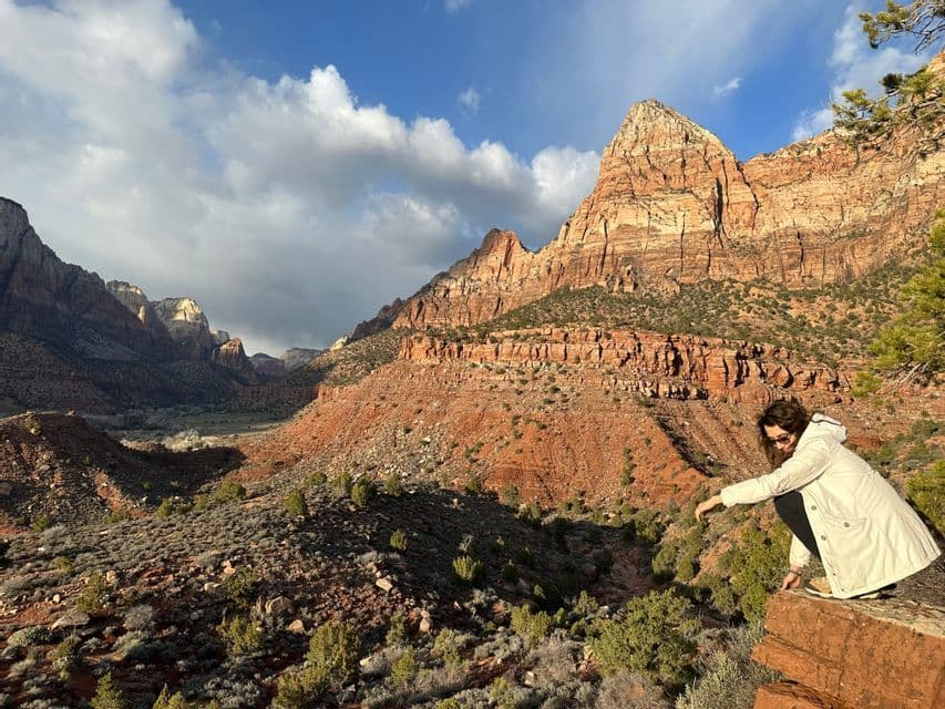 Una persona con chaqueta blanca se cuclilla en una cornisa rocosa, contemplando un vasto cañón de roca roja bajo un cielo parcialmente nublado.