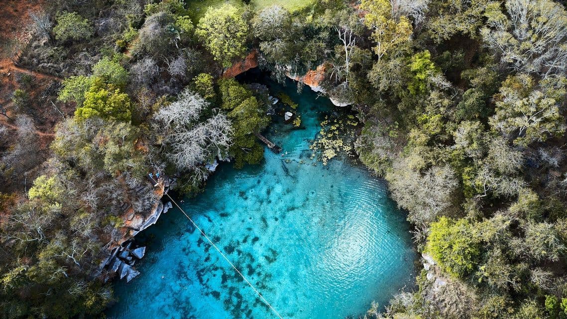 Una vista aerea dall'alto di un cenote turchese circondato da una fitta foresta, con una zipline che attraversa l'acqua limpida.