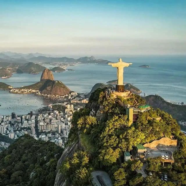 Una vista aerea della statua del Cristo Redentore su una montagna che domina una città costiera, una baia e altre montagne all'alba.
