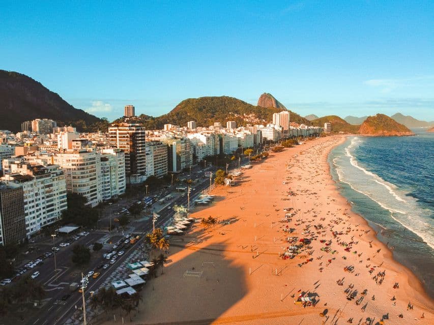 Vista aerea di una spiaggia sabbiosa affollata vicino a una città densa, con grandi montagne verdi sullo sfondo all'ora d'oro.